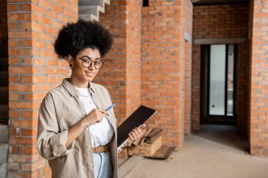 positive african american real estate agent with pen and folder looking at camera in new cottage clipart