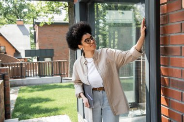 Mutlu Afro-Amerikan emlakçı. Dosyası modern kır evinin penceresine dokunuyor.