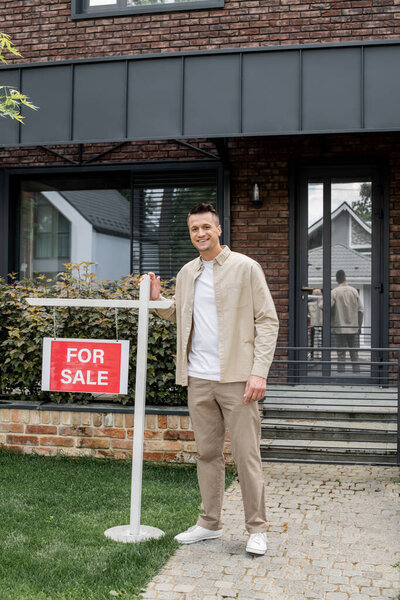 успешный и счастливый риэлтор недвижимости глядя на камеру рядом с современным коттеджем и на продажу буквы