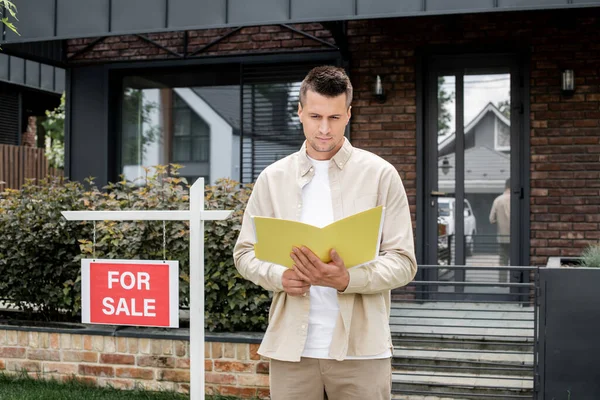 stock image confident real estate agent looking in folder near for sale signboard and modern cottage in city