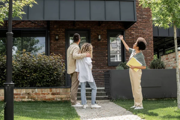 african american property agent pointing with hand near couple embracing next to modern cottage