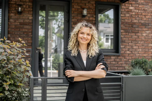 blonde and happy real estate agent standing with folded arms and looking at camera near cottage