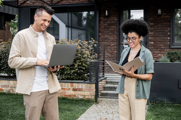 happy multiethnic real estate agents standing with laptop and folder near modern cottage
