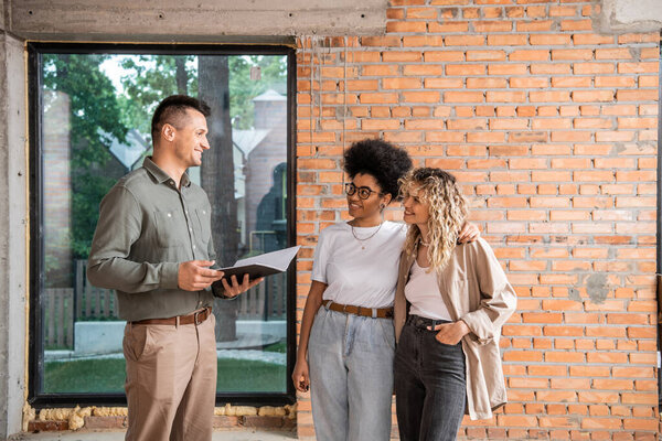 smiling real estate agent with folder talking to multiethnic lesbian couple inside new cottage