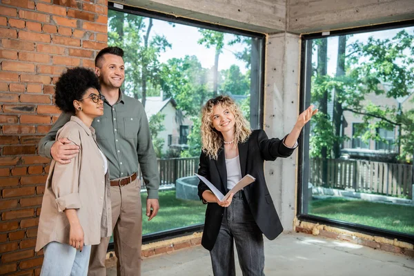 smiling real estate broker pointing with hand while showing new dwelling to multiethnic couple