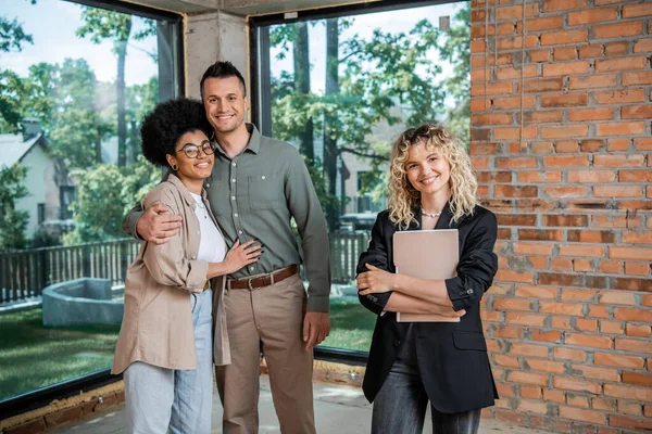 successful realtor with folder and cheerful multiethnic couple looking at camera in new cottage