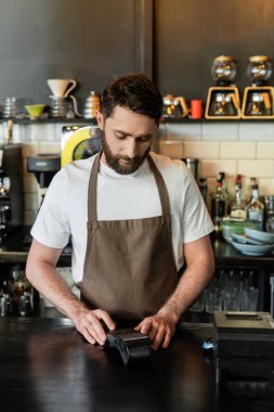 bearded barista in apron using payment terminal on bar while working in coffee shop clipart