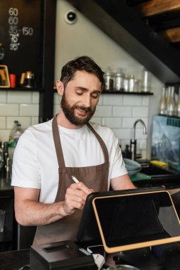 Kahve dükkanında çalışırken dijital tablet kullanan önlüklü gülümseyen sakallı barista.