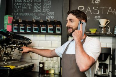 Kafede kahve makinesinin yanında çalışırken önlüklü gülümseyen bir barista akıllı telefondan konuşuyor.