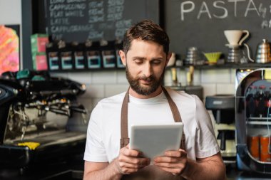 Kafede çalışırken ve barın yanında dururken dijital tablet kullanan önlüklü bir barmen.