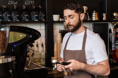 bearded barista in apron holding brush and coffee machine holder while working in coffee shop clipart