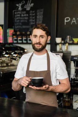 Önlük takmış, elinde kahve fincanı tutan ve kahve dükkanında kameraya bakan sakallı bir barista.