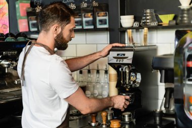 Kafe 'de çalışırken kahve makinesinin yanındaki tutacağa kahve dolduran önlüklü baristanın yan görüntüsü