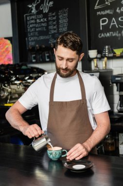 bearded barista in apron pouring milk from pitcher while making cappuccino on bar in coffee shop clipart