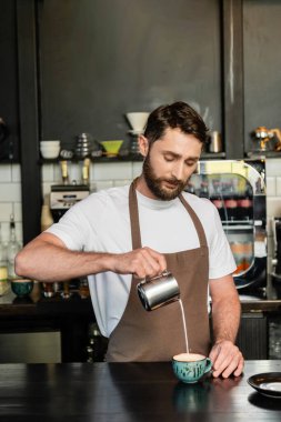 Önlüklü sakallı barista kahve dükkanında kapuçino içerken sütten süt dolduruyor.