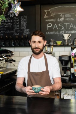 Önlüklü gülümseyen barista kameraya bakıyor, elinde bir fincan kapuçino tutuyor ve kahve dükkanında çalışıyor.