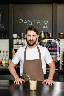 Önlüklü neşeli barista, kahve dükkanının barında, soğuk kahve bardağının yanında kameraya bakıyor.