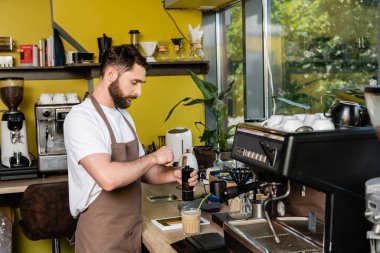 Önlüklü barista dijital tabletin yanında kahve öğütüyor ve kafede soğuk kahve içiyor.