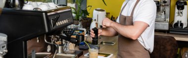Önlüklü barista, dijital tabletin yanında kahve öğütüyor ve kafede soğuk kahve içiyor.
