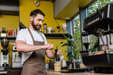 Önlüklü gülümseyen barista kahve öğütüyor atölyedeki soğuk içkinin yanında.