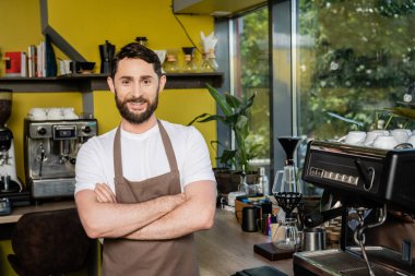 Önlüklü gülümseyen barista kollarını kavuşturur ve kahve makinesinin yanındaki kameraya bakar.