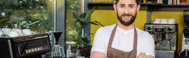 Kafede kahve makinesinin yanında afiş asarken kameraya bakan önlüklü neşeli barista.