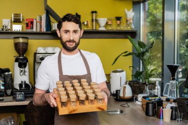 smiling barista in apron holding jars with coffee beans and looking at camera in coffee shop clipart