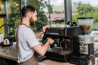 Kafede kahve makinesiyle çalışan önlüklü sakallı baristanın yan görüntüsü.