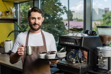 Önlüklü güler yüzlü barista kahve makinesinin yanında sürahi ve fincanlar tutuyor.
