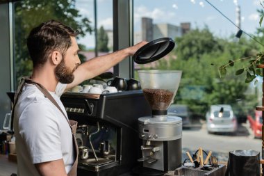 Önlüklü baristanın kahve öğütücüsü kullanırken arka planda kahve dükkanında çalışmasının yan görüntüsü