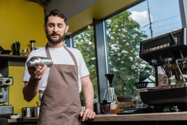 bearded barista in apron using shaker while working near coffee machine in coffee shop clipart