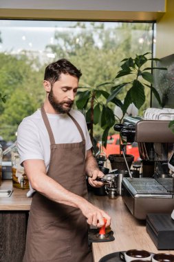 Önlüklü sakallı barista kahve makinesinin yanında kahve makinesi tutuyor.