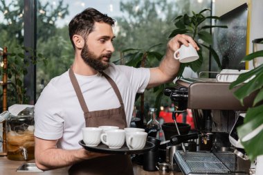Kafede çalışırken önlüklü sakallı barista kahve makinesine temiz bardaklar koyuyor.