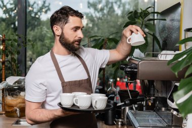 bearded barista in apron putting clean cups on coffee machine while working in coffee shop clipart