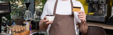 cropped view of barista in apron holding credit card and cup while working in coffee shop, banner