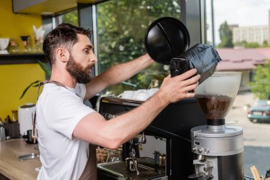 side view of barista in apron pouring coffee beans in grinder while working in coffee shop clipart