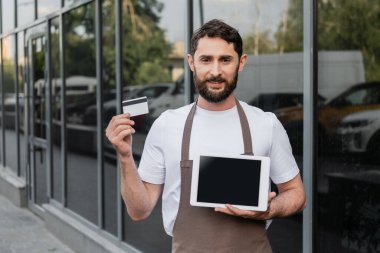 smiling barista holding credit card and digital tablet with blank screen on urban street