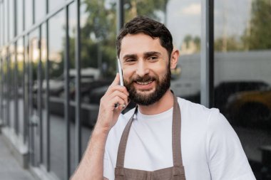 Önlüklü neşeli barista akıllı telefonla konuşuyor ve arka planda şehir sokağında duruyor.