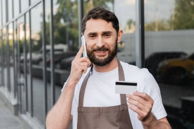 Elinde kredi kartı olan neşeli barista ve arka plandaki şehir caddesinde akıllı telefondan konuşuyor.