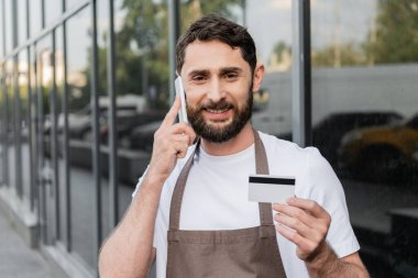 cheerful barista holding credit card and talking on smartphone on urban street on background