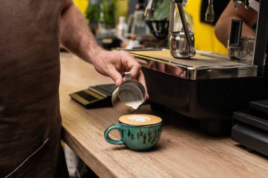 Kafede kapuçino içmek için kahve bardağındaki sürahiden süt döken önlüklü barista manzarası.