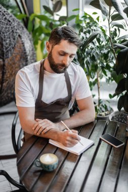 Önlüklü barista, akıllı telefon ve kahve dükkanındaki masada bir fincan kapuçino yanında deftere yazıyor.