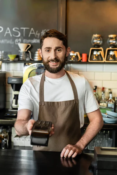 smiling barista in apron holding payment terminal and looking at camera in coffee shop