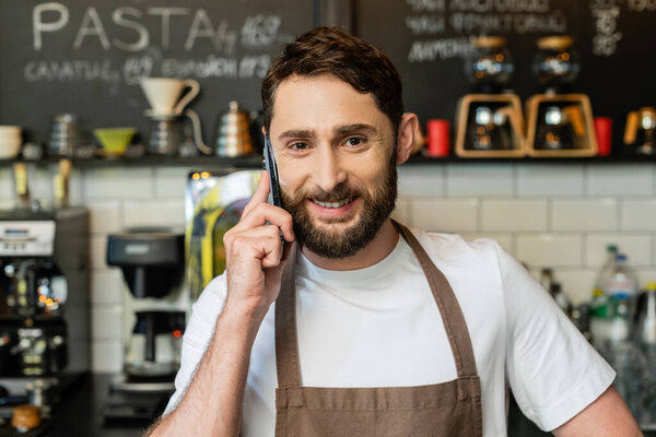 smiling bearded barista in apron talking on smartphone and looking at camera in coffee shop