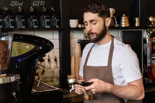コーヒーショップで働いている間 ブラシおよびコーヒーマシンのホールダーを握るエプロンのひげ付きのバリスタ — ストック写真