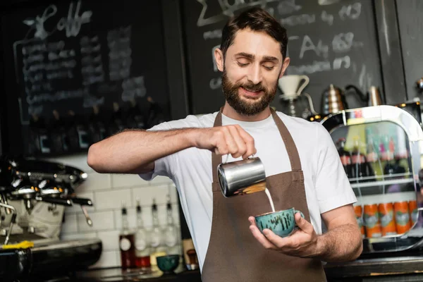 stock image positive barista in apron pouring milk from pitcher in cup with cappuccino in coffee shop