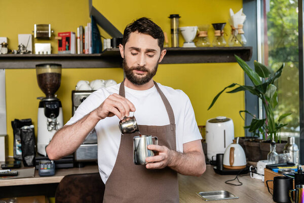 бородатый бариста в фартуке наливая кофе в металлический кувшин во время работы в кафе на заднем плане