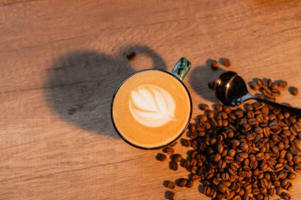 top view of cup of cappuccino near coffee beans and spoon on worktop in coffee shop