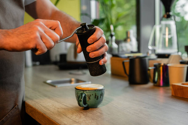 cropped view of barista grinding cinnamon above cup of cappuccino on worktop in coffee shop