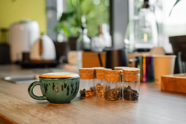 cup of cappuccino near coffee beans in jars on worktop in blurred coffee shop